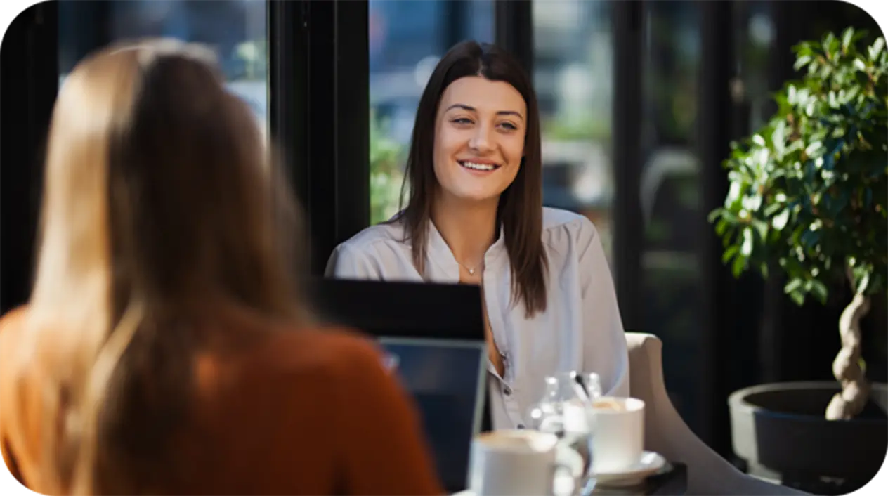 Two women having a coaching conversation at a sunlit café table, one smiling and engaged while the other listens attentively — representing supportive, purpose-driven career coaching